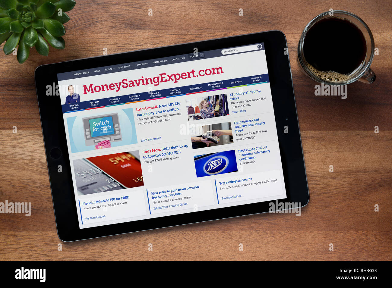 Il sito web di MoneySavingExpert è visto su un tablet iPad, su di un tavolo di legno lungo con un caffè espresso e una pianta di casa (solo uso editoriale). Foto Stock