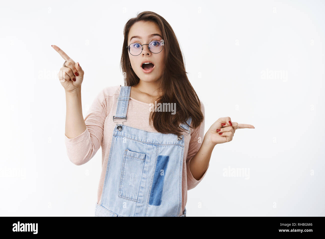 Curioso e smart giovani creativi 20s europea femminile in bicchieri e tuta in jeans a bocca aperta interrogato e colpito come puntamento lateralmente incuriosito Foto Stock