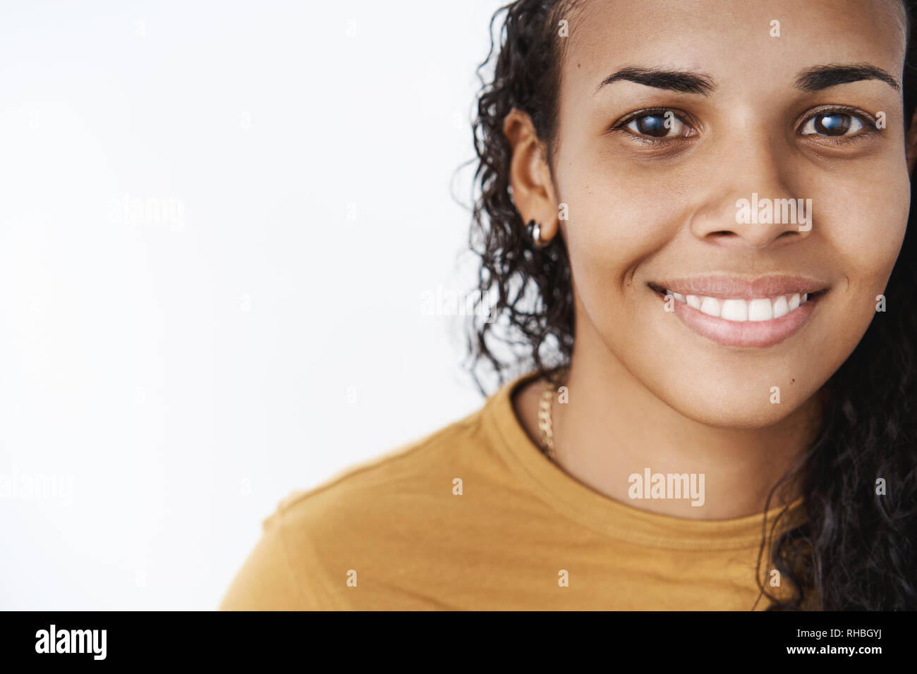 Close-up shot di sorridere felice e soddisfatto amichevole-cercando AFRICAN-AMERICAN donna con capelli ricci in senape t-shirt grinning e ammirate la fotocamera Foto Stock