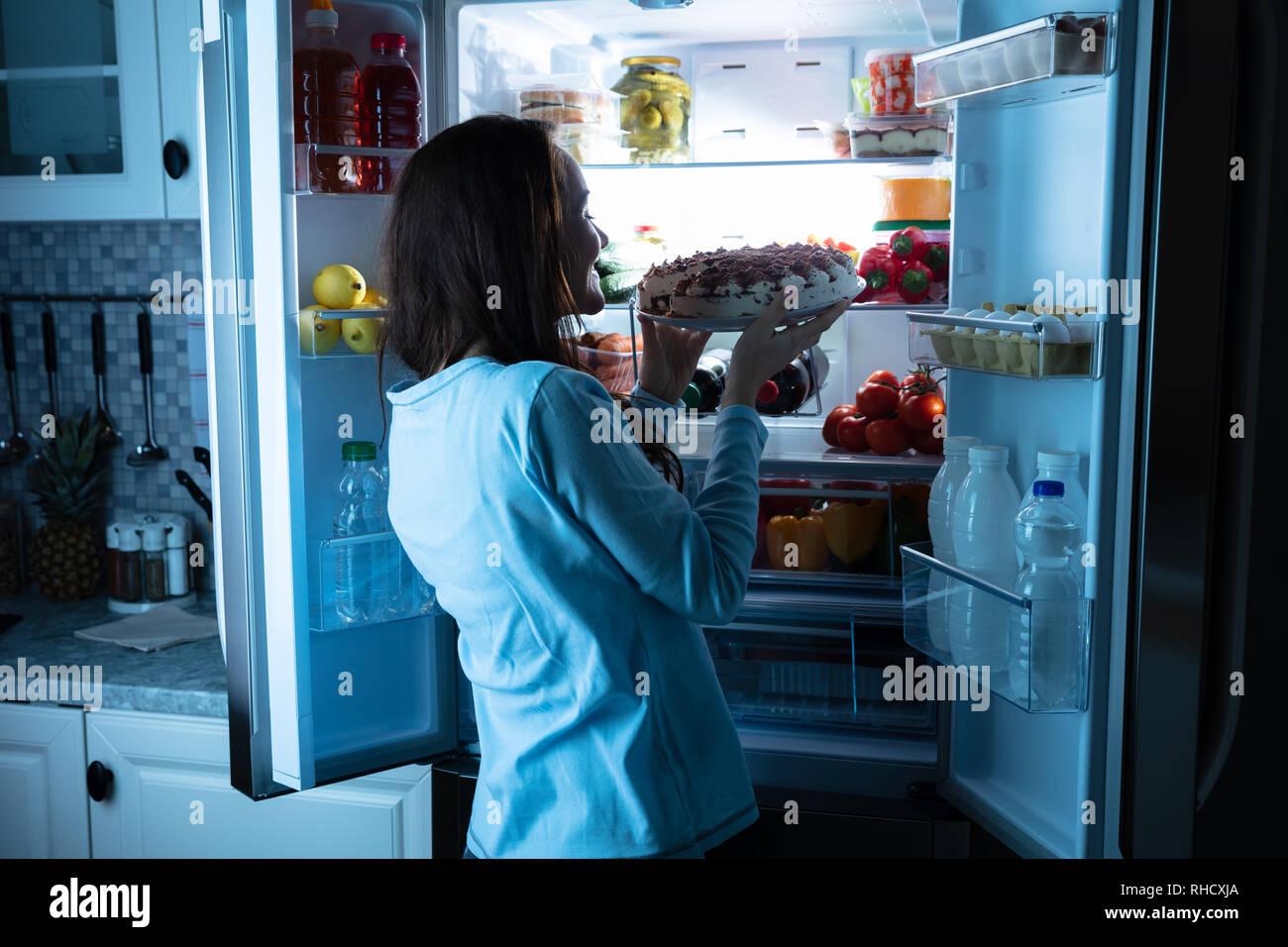 Sorridente giovane donna che guarda la torta nella parte anteriore di un frigorifero aperto Foto Stock