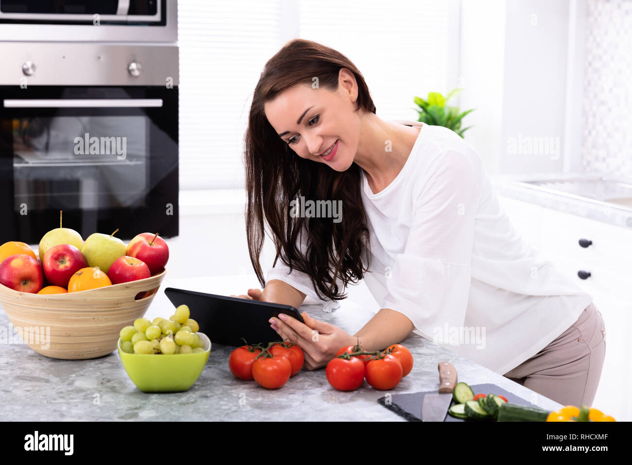 Felice giovane donna con tavoletta digitale nei pressi di verdure fresche sul banco di cucina Foto Stock