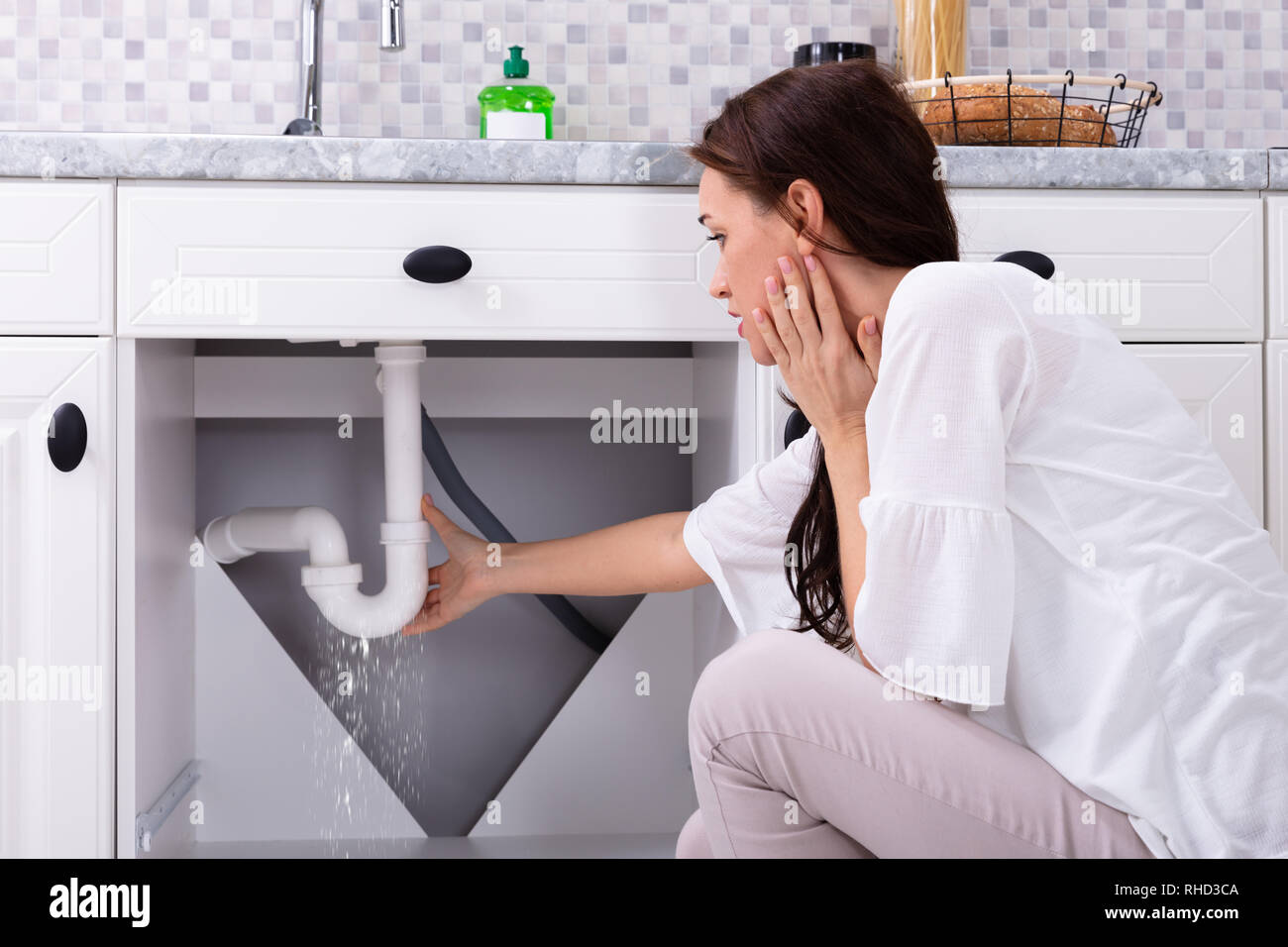 Vista posteriore di una donna che cerca di fermare la perdita di acqua dal tubo di lavello in cucina Foto Stock