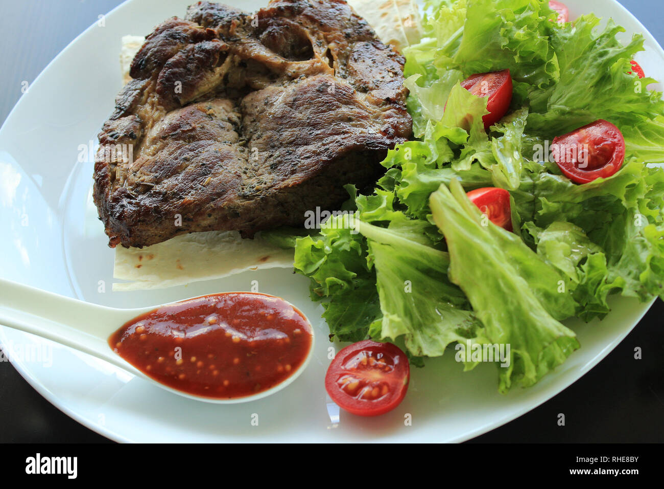 La carne fritta di filetto con verdi, salsa piccante e pomodori ciliegia in piastra bianca sul tavolo di legno isolato Foto Stock