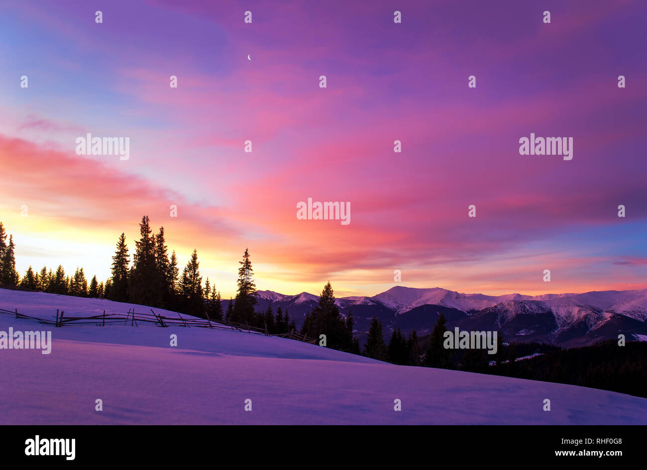 Inverno mattina paesaggio. Il sole sorge alle spalle di alberi e illumina le nuvole. Montagna cresta. La luna nel cielo. Recinzione in legno in campo nevoso. Twili Foto Stock