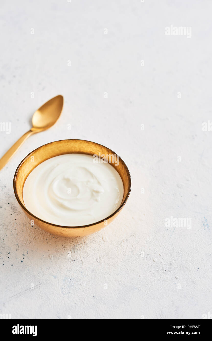 Ciotola di fresco lo yogurt greco e il cucchiaio d'oro bianco su sfondo astratto. Una sana prima colazione. Golden utensili. Vista dall'alto. Copia spazio per il testo. Foto Stock