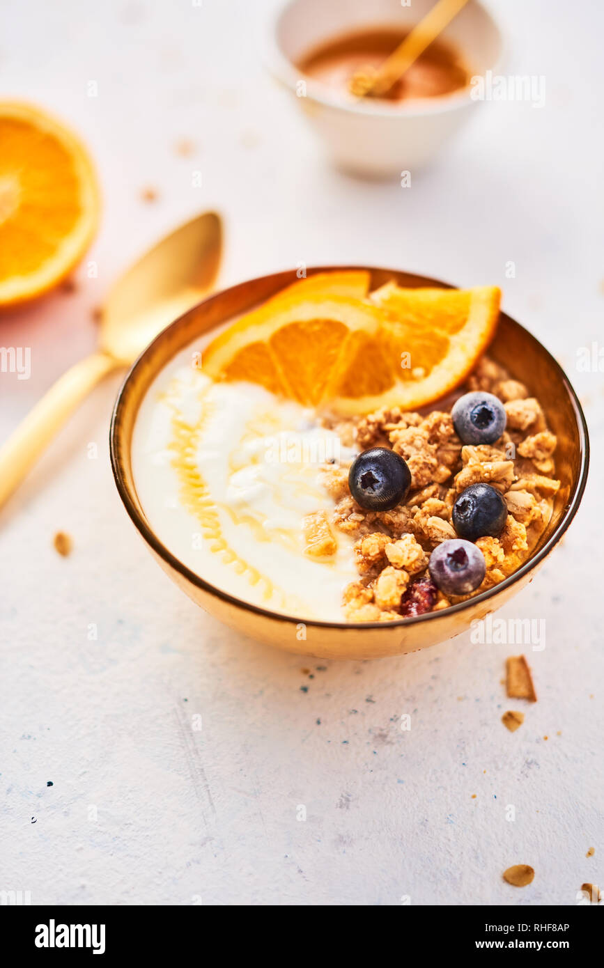 Ciotola di muesli fatti in casa con yogurt, miele, succo di arancia e mirtilli surgelati bianco su sfondo astratto. Golden utensili. Vista dall'alto. Copia spazio f Foto Stock