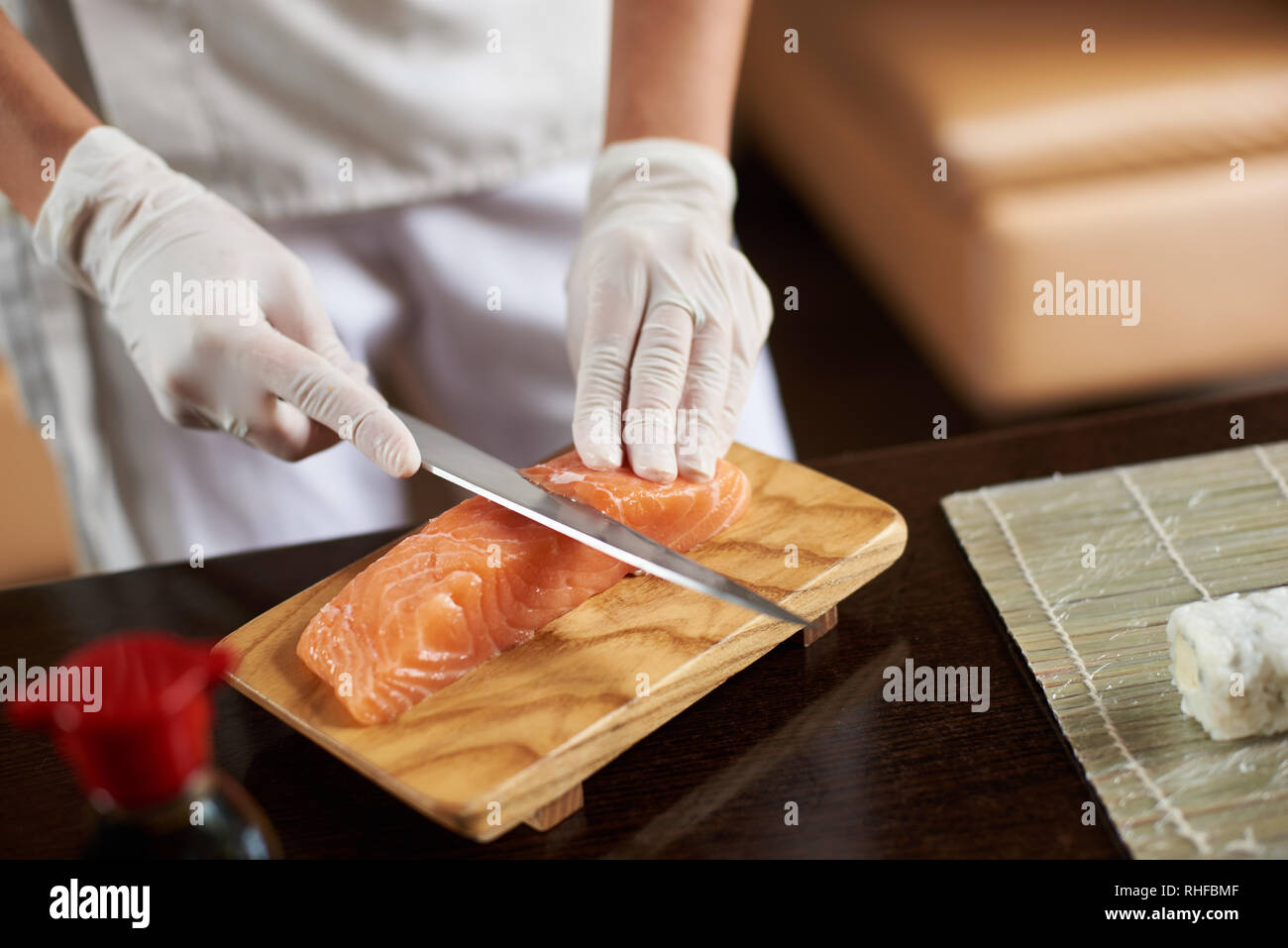 Vista da vicino il processo di preparazione del sushi di laminazione. Mani in guanti monouso per affettare il salmone sul pannello di legno Foto Stock