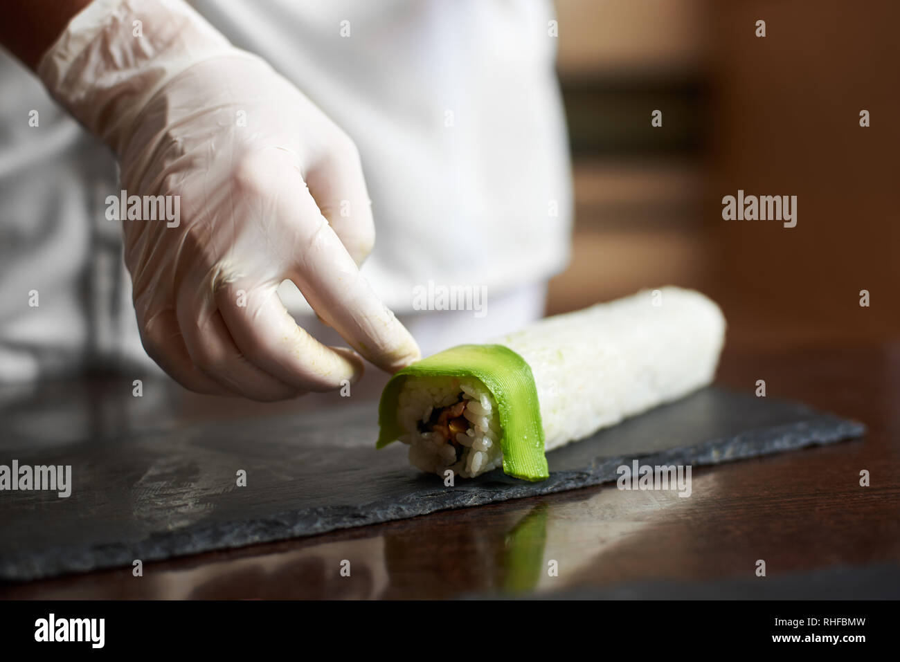 Vista da vicino il processo di preparazione del sushi di rotolamento a. Femmina in mano guanto decora il rotolo con fette di avocado Foto Stock