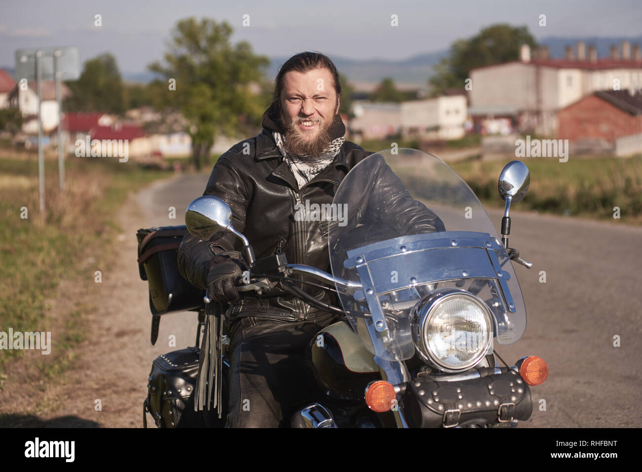 Ritratto di bello sorridenti barbuto biker in black leather jacket seduto sul motociclo moderno sulla strada del paese su sfondo sfocato di piccola città edifici sulla soleggiata giornata estiva. Foto Stock
