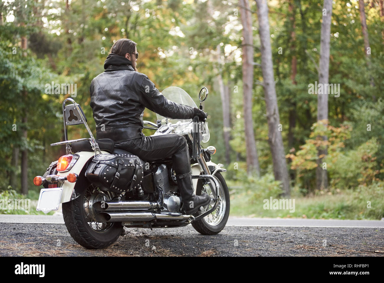 Vista posteriore del barbuto biker in pelle nera abbigliamento equitazione sulla cruiser moto sulla strada del paese su sfondo di vuoto dritta strada asfaltata e verdi alberi bokeh fogliame. Foto Stock