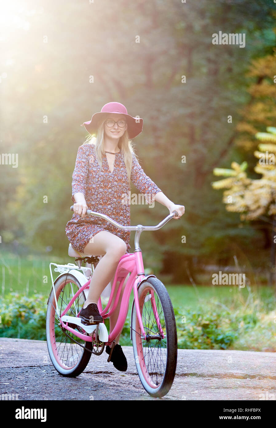 Slim sorridenti bionda di moda donna attraente in bicchieri, abito corto e Red Hat lady di equitazione Bicicletta lungo lastricata park alley sul bel verde e alberi d'oro illuminata dal sole luminoso dello sfondo. Foto Stock