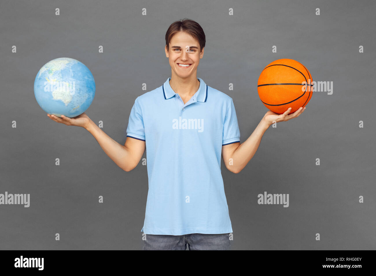 Giovane uomo in piedi isolato sul muro grigio azienda globo terrestre e basket ball cercando fotocamera a sorridere allegro Foto Stock