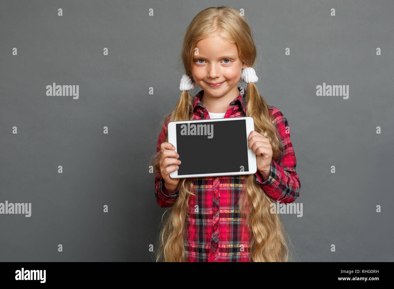 Bambina con due ponytails studio isolato permanente sulla parete grigia mostra schermo della tavoletta digitale cercando fotocamera a sorridere giocoso Foto Stock