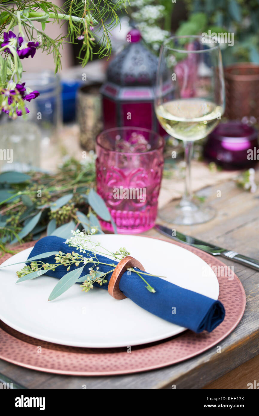 Posizionare l'impostazione sul cortile esterno dinner party tabella- piastra bianca su rame caricabatterie con blue igienico in anello, con fiori, candele, vetro e vino. Foto Stock
