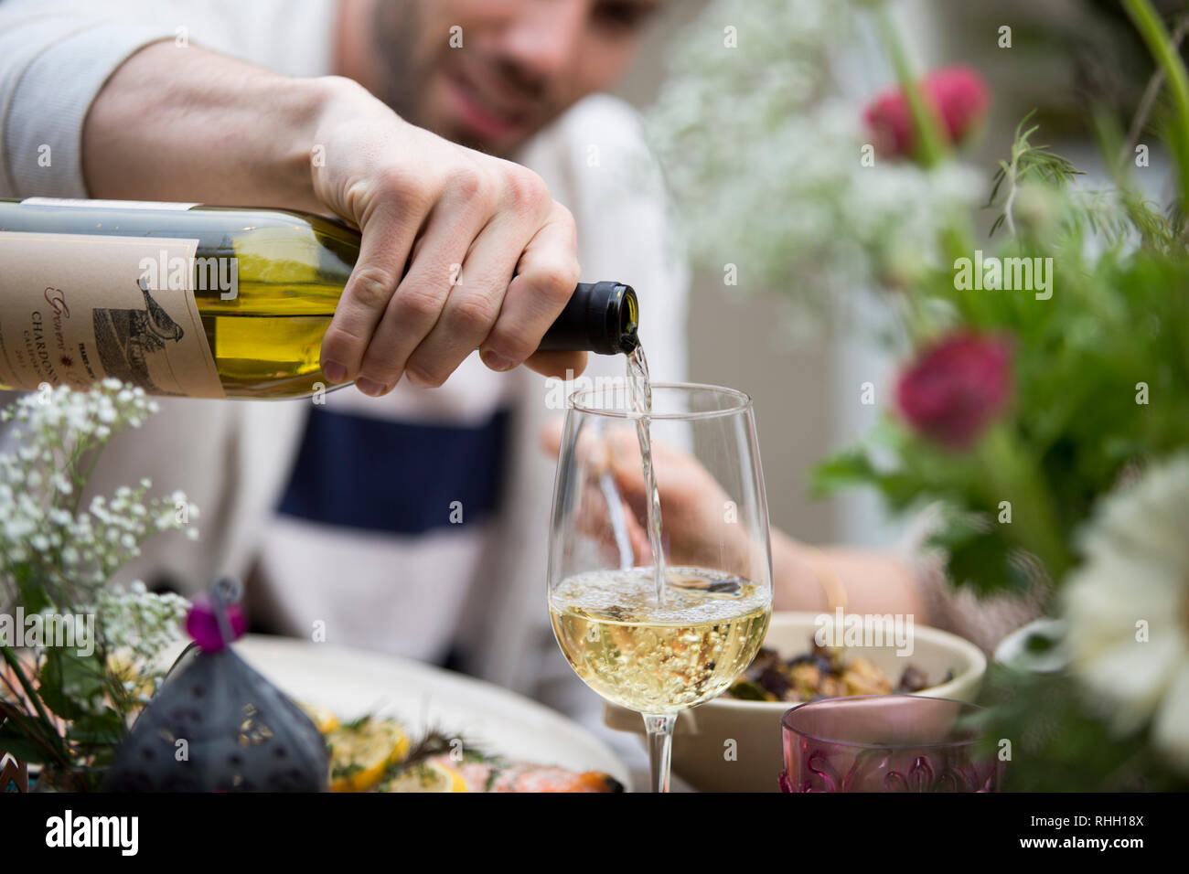 Mano maschio versa del vino bianco in vetro al picnic esterno dinner party tavolo con fiori, volto sfocato in background. Foto Stock