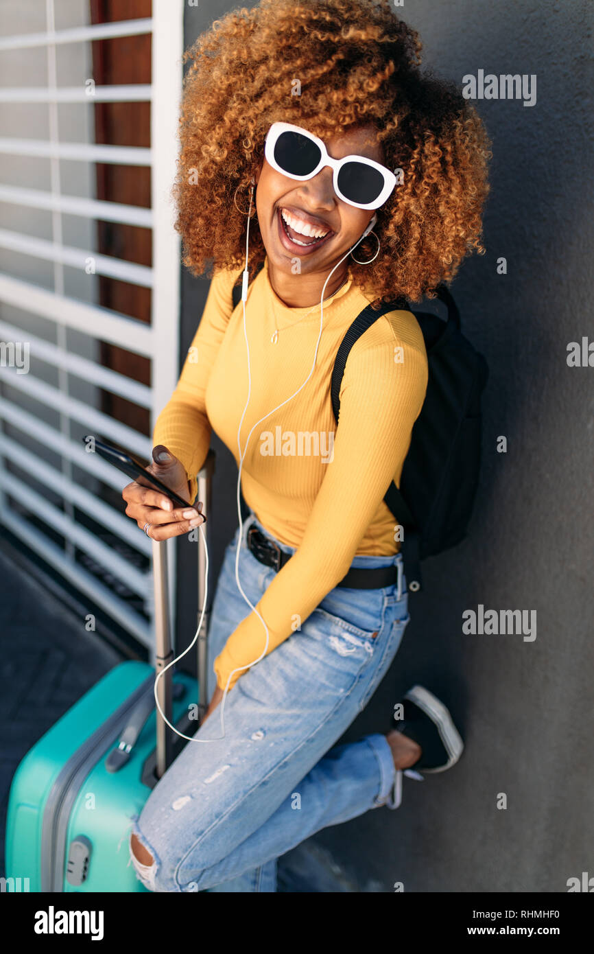 Donna sorridente in piedi contro un muro mentre tiene il suo sacchetto di bagagli e telefono cellulare. Afro American tourist donna in occhiali da sole in piedi con i suoi bagagli li Foto Stock