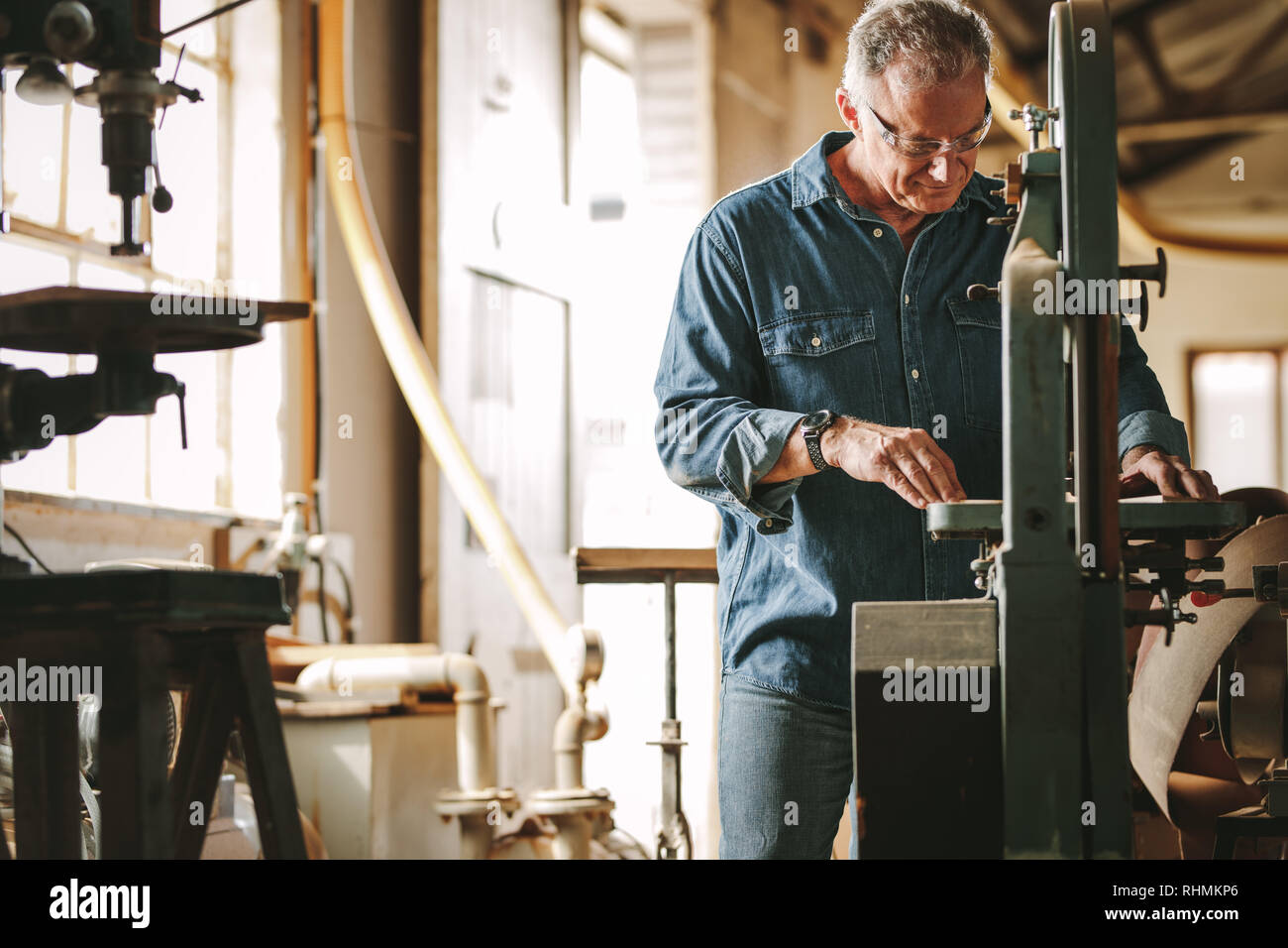 Maschio maturo falegname lavorando su una sega a nastro macchina nel laboratorio di falegnameria. Senior uomo taglio del legno sulla macchina. Foto Stock