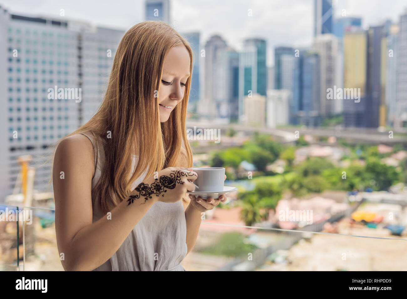 Giovane donna con una foto della mano di henna - mehendi beve caffè al mattino sul balcone che si affaccia sulla grande città e grattacieli Foto Stock