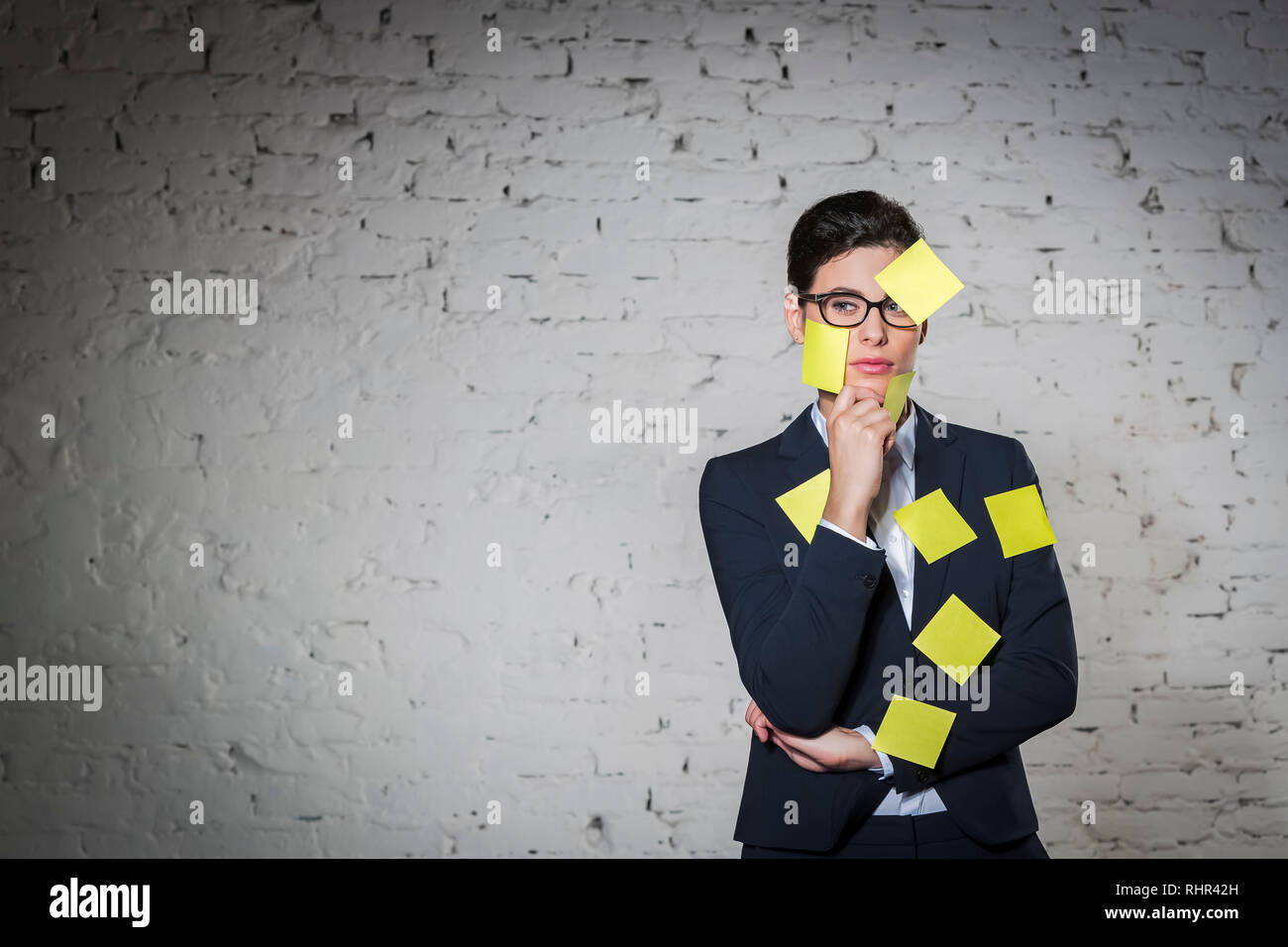 Riflessivo imprenditrice con giallo vuota note adesive sulla faccia e tuta contro la parete in ufficio Foto Stock