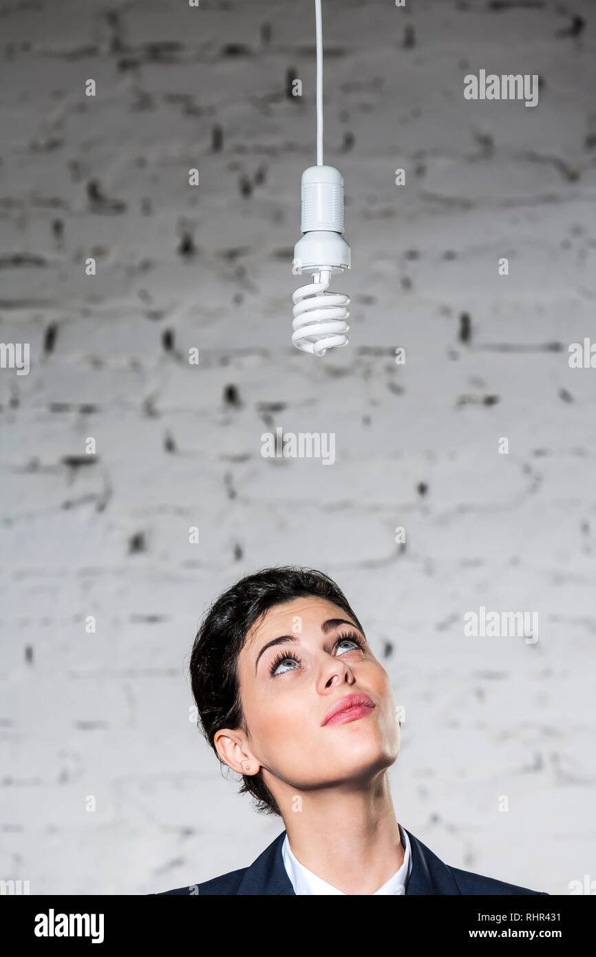 Giovane imprenditrice pensieroso guardando lampadina a basso consumo appeso contro un muro di mattoni in ufficio Foto Stock