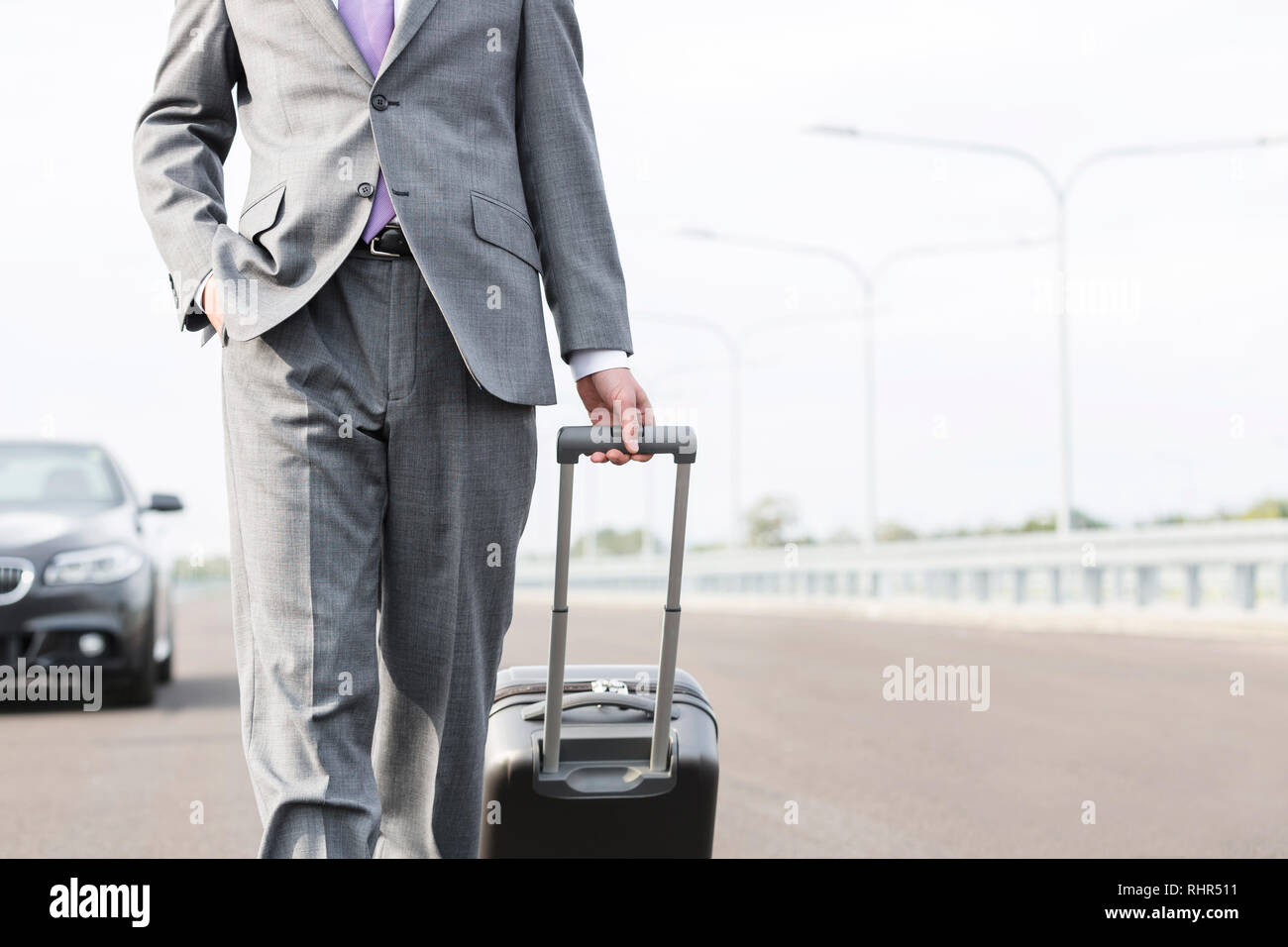 Sezione mediana di imprenditore a piedi con la valigia sulla strada contro sky Foto Stock