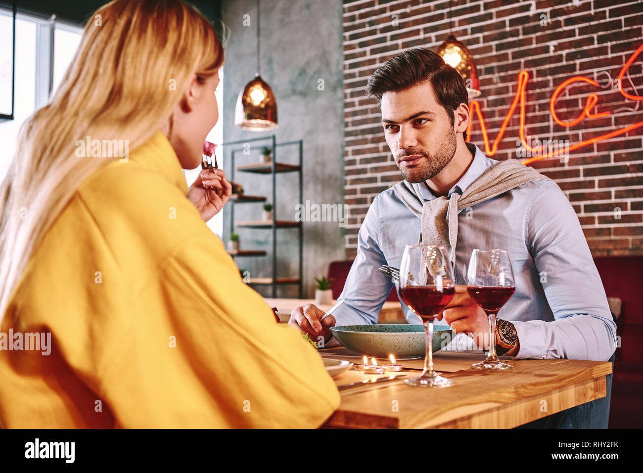 Cena romantica. Uomo in camicia blu mangiare e parlare con la fidanzata. Rose rosse sono sdraiato sul tavolo. Giovane donna bionda in maglione di senape Foto Stock