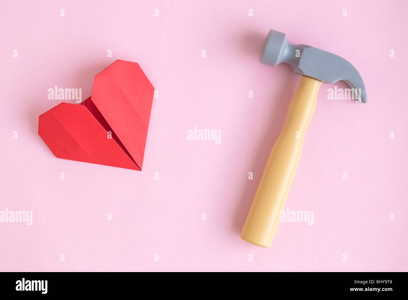 Piatto di laici con un martello di plastica e carta origami cuore contro di rosa pastello minima di sfondo heartbreak concetto creativo. Foto Stock