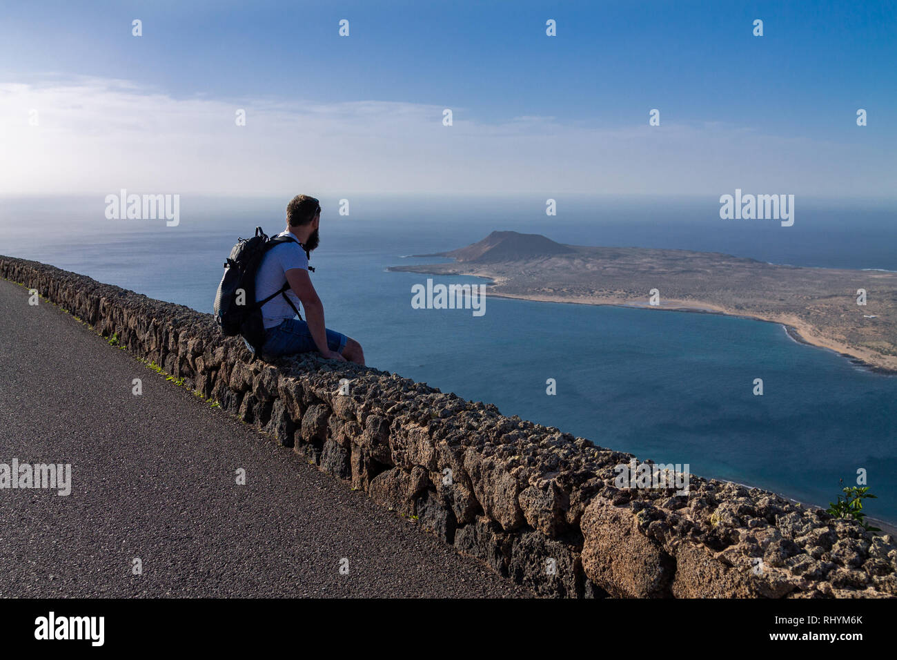 Un ragazzo in abiti estivi è seduta su una pietra recinzione e guarda alla vicina isola nell'oceano. Mirador del Rio Lanzarote, Spagna. Foto Stock