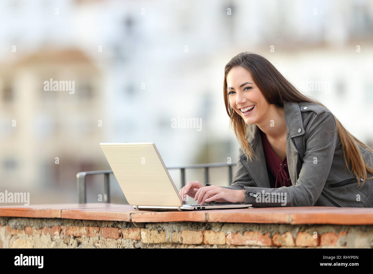 Donna felice con un computer portatile e guardando a voi in un balcone in una città in periferia Foto Stock