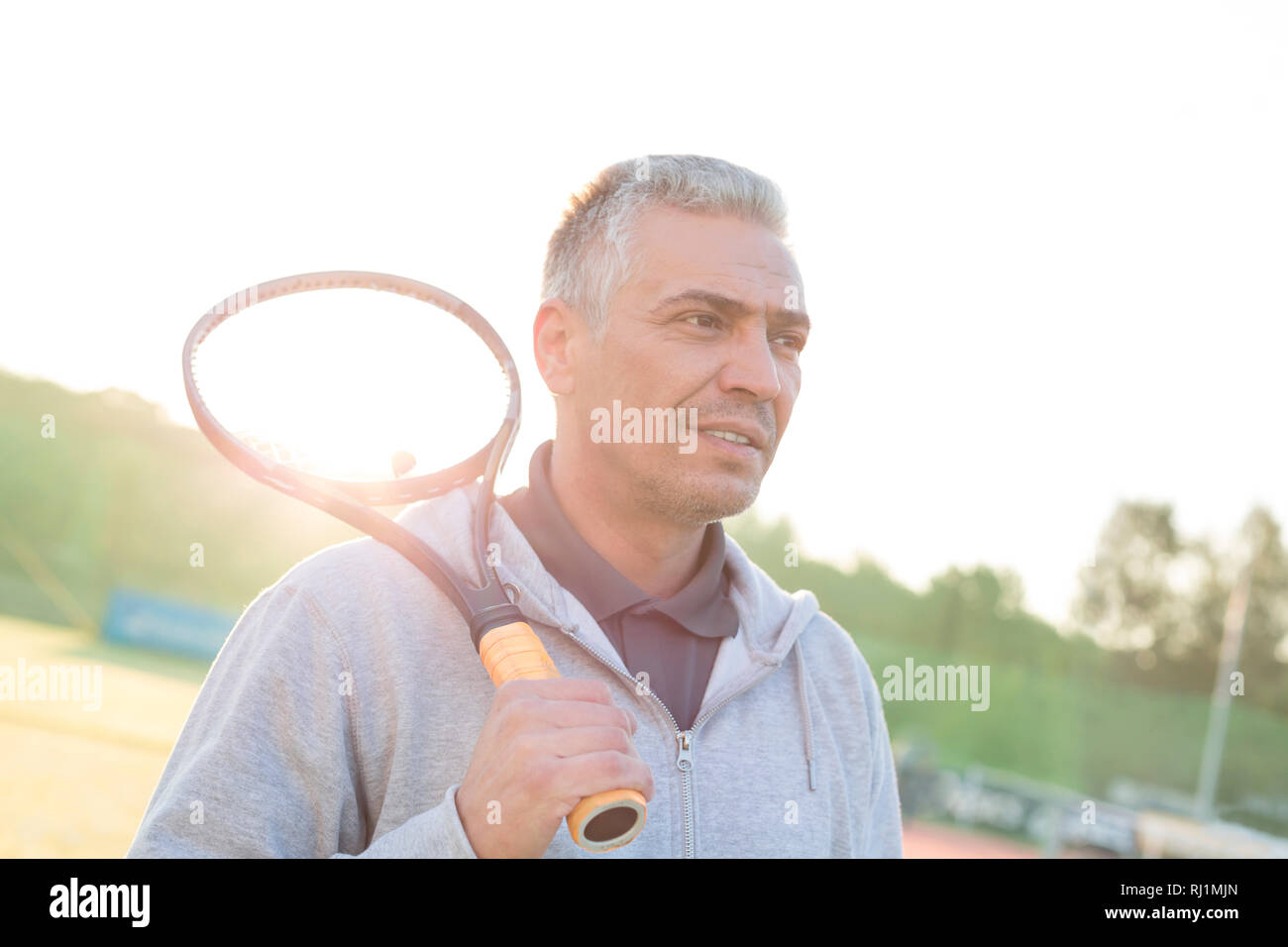 Back lit di fiducioso uomo maturo in piedi con la racchetta da tennis in tribunale contro il cielo chiaro Foto Stock