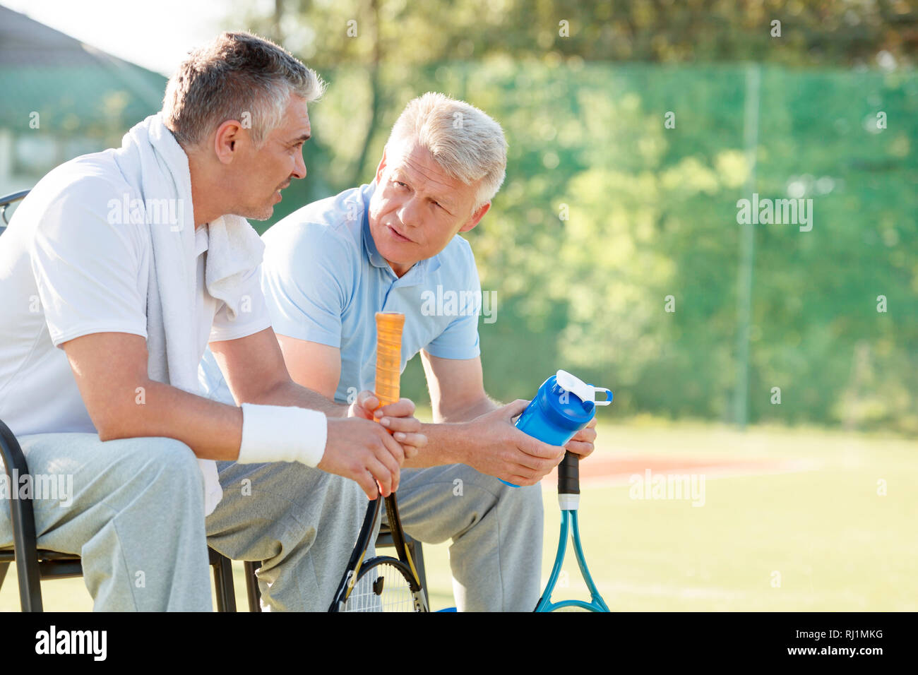Maschio maturo amici parlare mentre si siede con racchette da tennis e una bottiglia d'acqua sulla sedia a corte Foto Stock