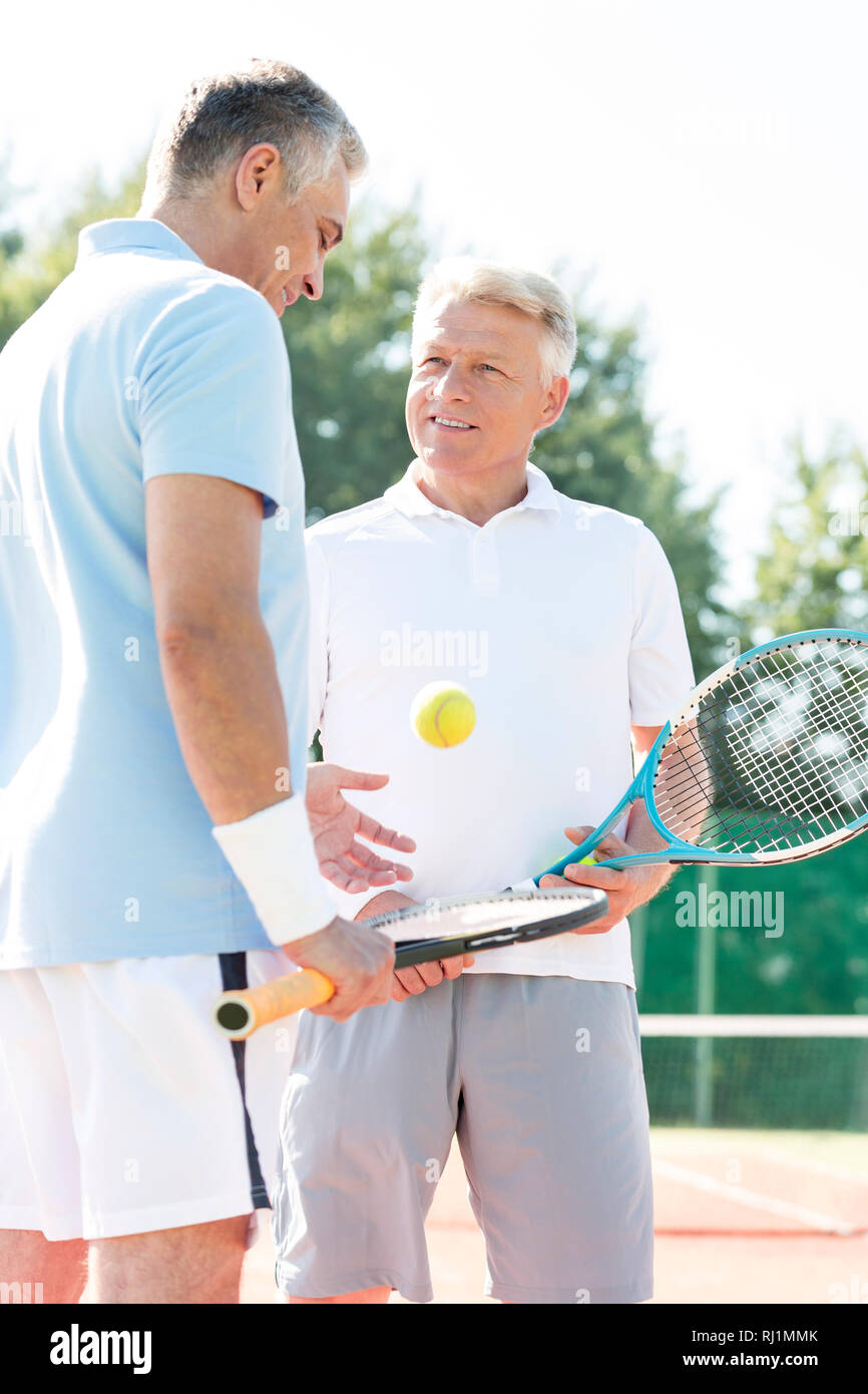 Gli uomini di età matura a parlare mentre in piedi con la racchetta e la palla sul campo da tennis durante il fine settimana estivo Foto Stock