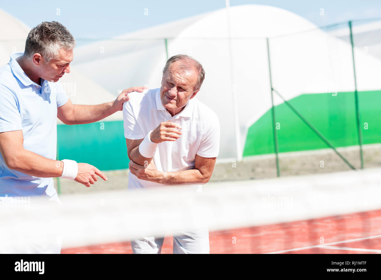 Uomo maturo aiutando senior amico con il gomito lesioni mentre gioca a tennis sulla giornata di sole Foto Stock
