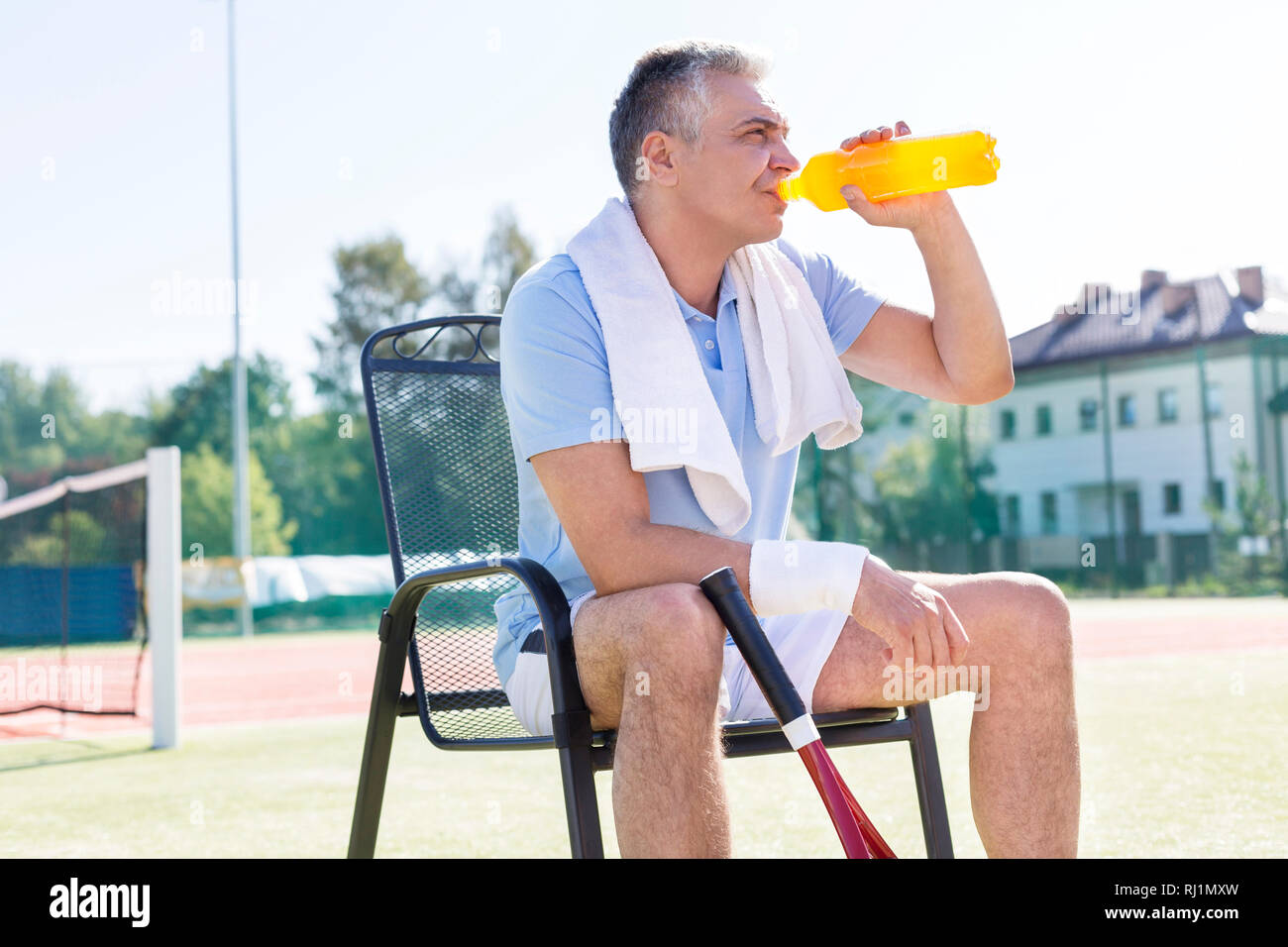 Stanco uomo maturo di bere dalla bottiglia mentre è seduto su una sedia al campo da tennis sulla giornata di sole Foto Stock