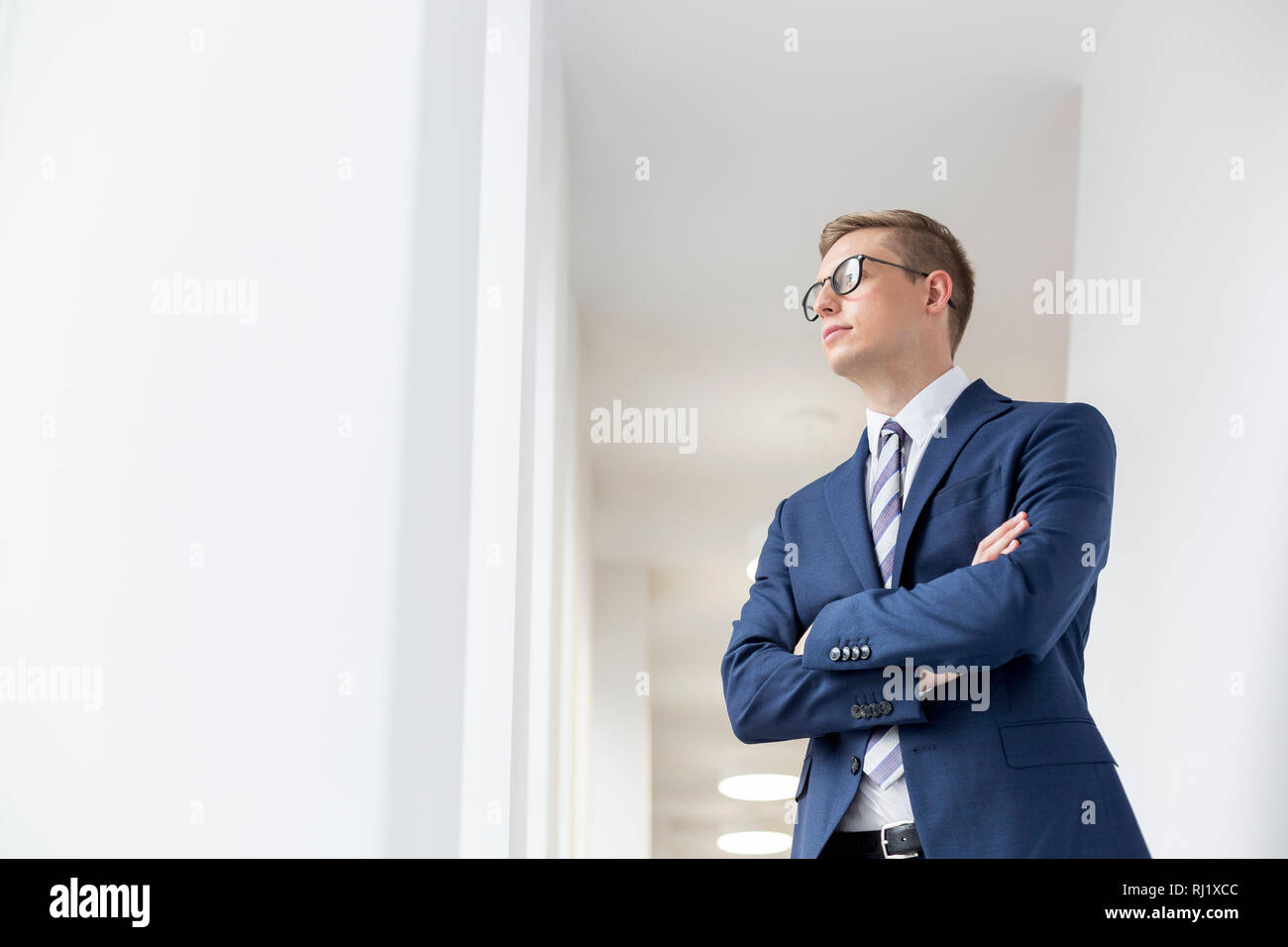 Giovane imprenditore pensieroso in piedi con le braccia incrociate che guarda lontano nel corridoio di office Foto Stock
