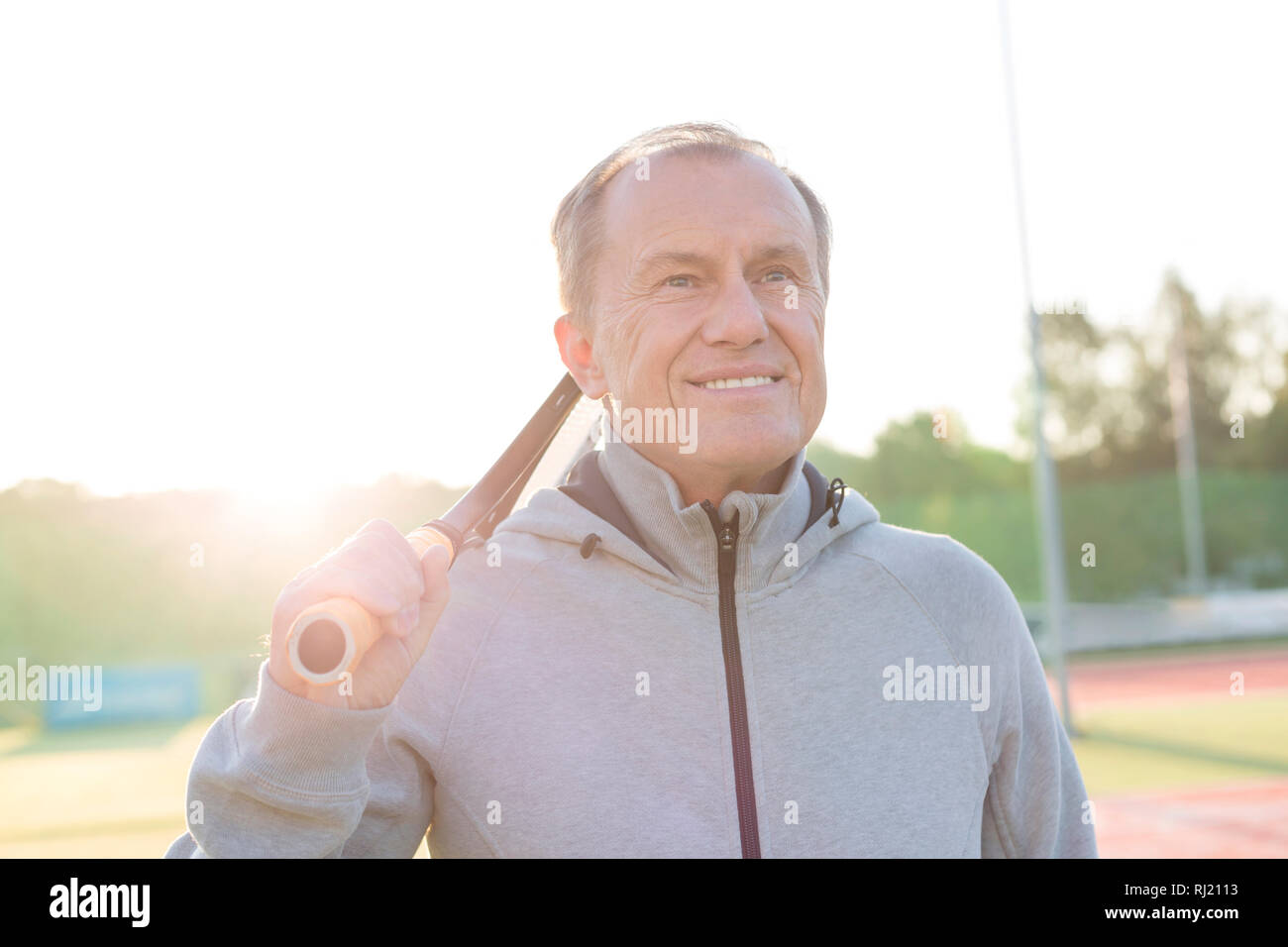 Senior sorridente uomo in piedi con la racchetta da tennis in tribunale contro il cielo chiaro Foto Stock