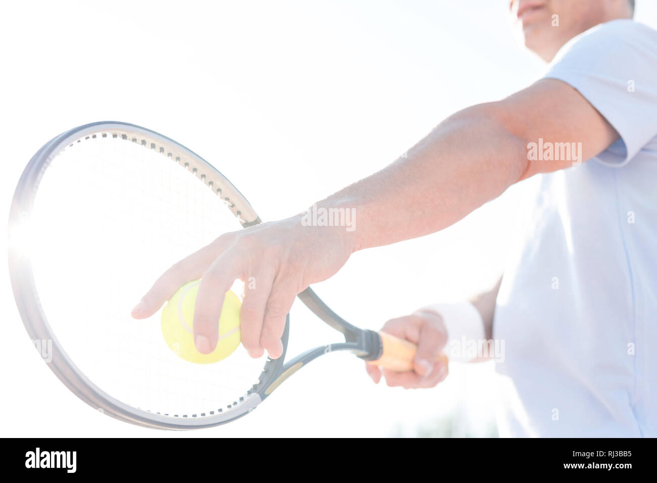 Sezione mediana di uomo maturo che serve palla da tennis contro il cielo chiaro sulla giornata di sole Foto Stock
