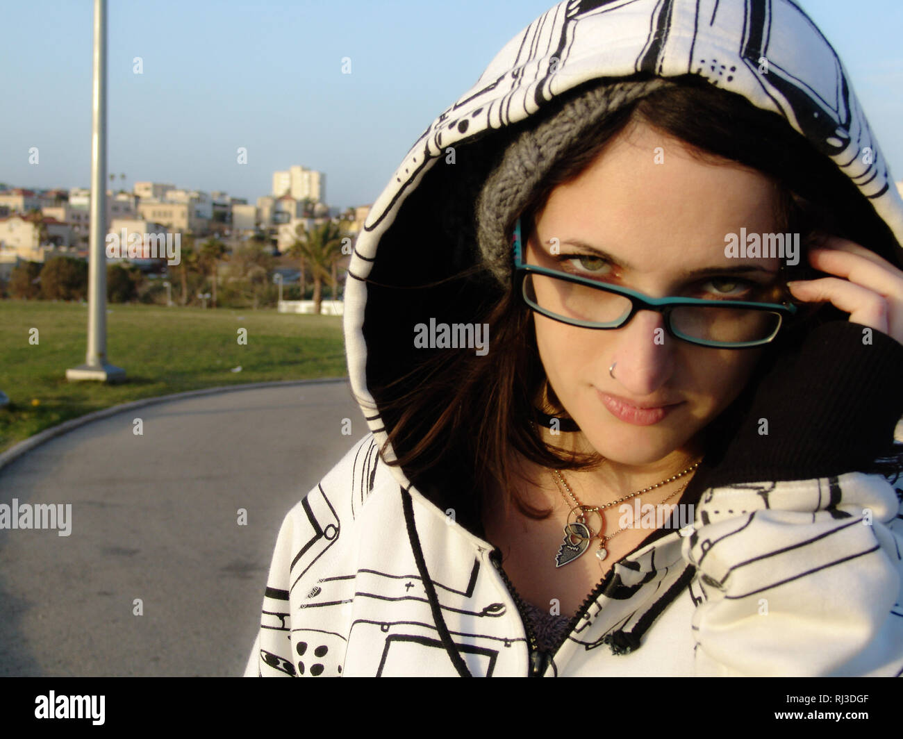 Giovane donna con verde blu occhi e occhiali, guardando la telecamera con un espressione seria in prima linea di un parco cittadino con complessi edilizi Foto Stock