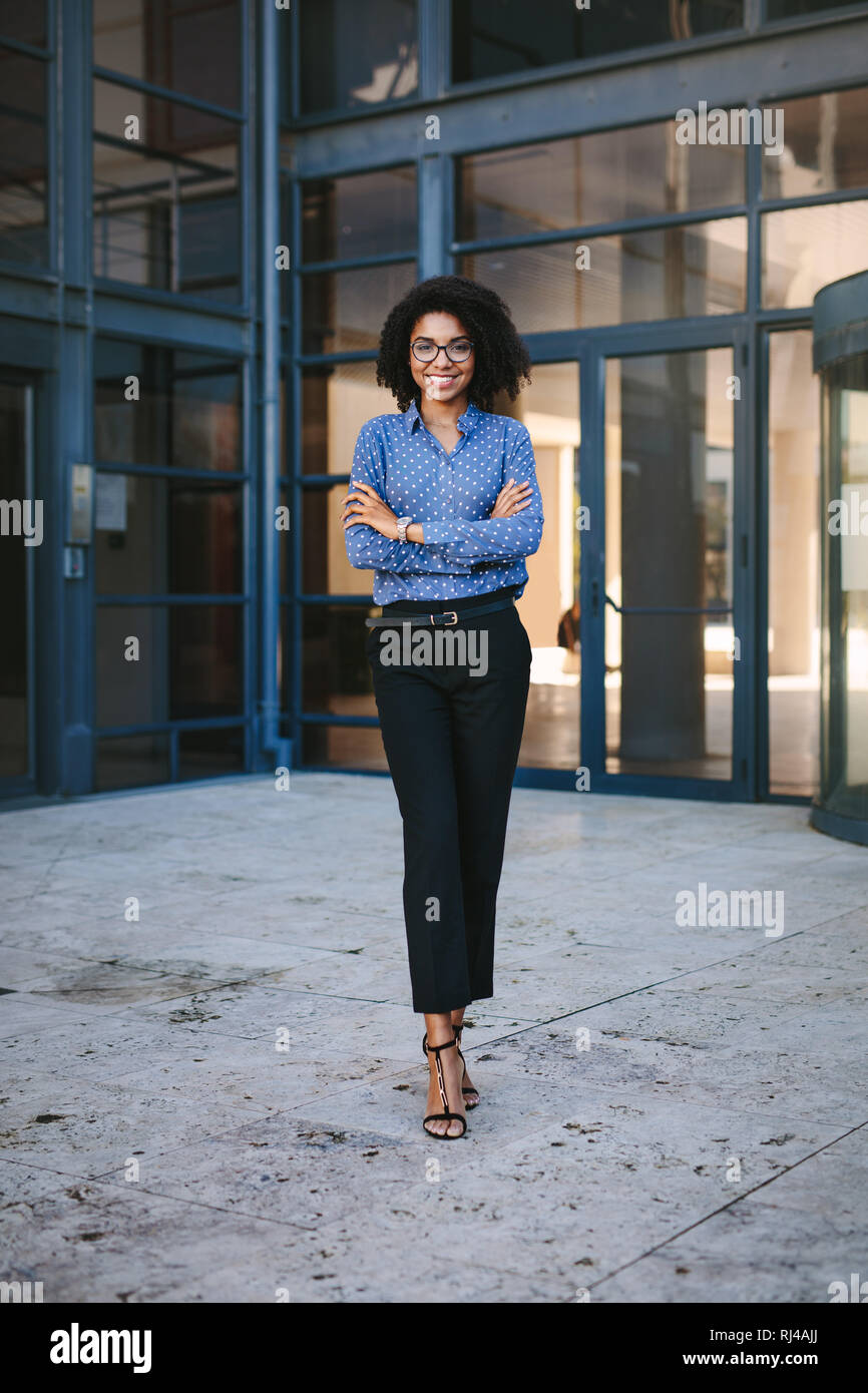 A piena lunghezza Ritratto di fiducioso giovane imprenditrice africana in piedi con le braccia incrociate. Donna in abiti formali guardando la fotocamera e sorridente. Foto Stock