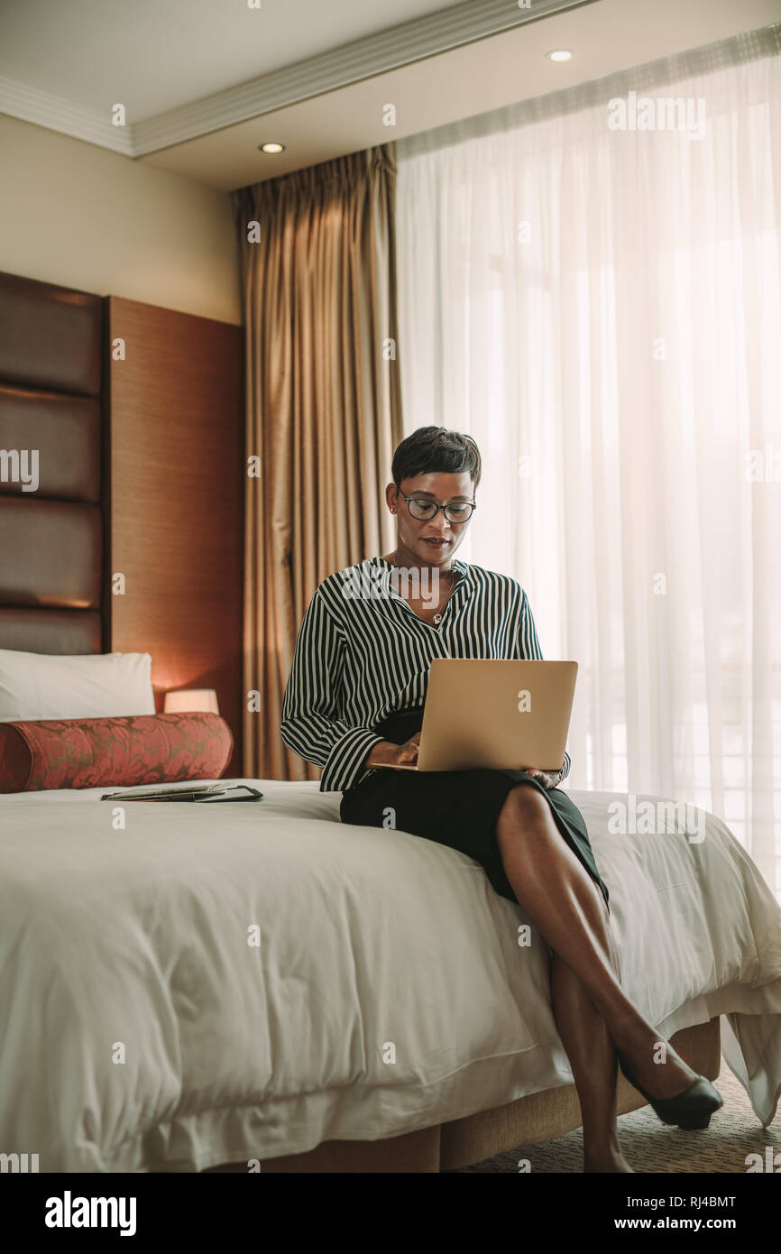 Donna africana seduta sul letto in camera in hotel utilizzando laptop. Business donna lavora da camera in hotel. Foto Stock