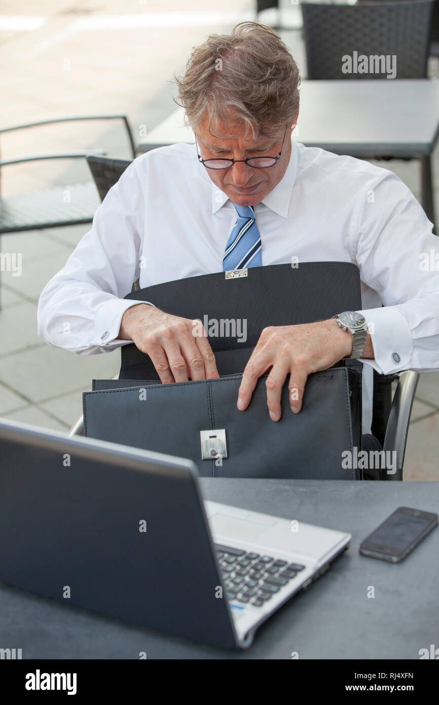 Geschäftsmann mit Laptop sitzt am Tisch in Straßenrestaurant und kramt in Aktentasche Foto Stock