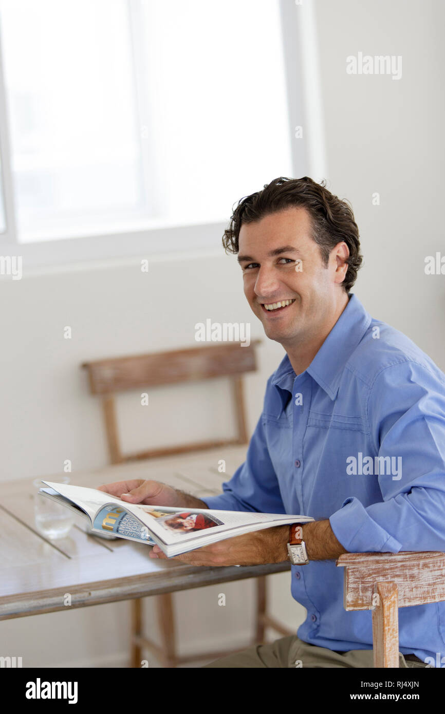 Mann sitzt am Tisch mit Laptop und blättert in Magazin Foto Stock