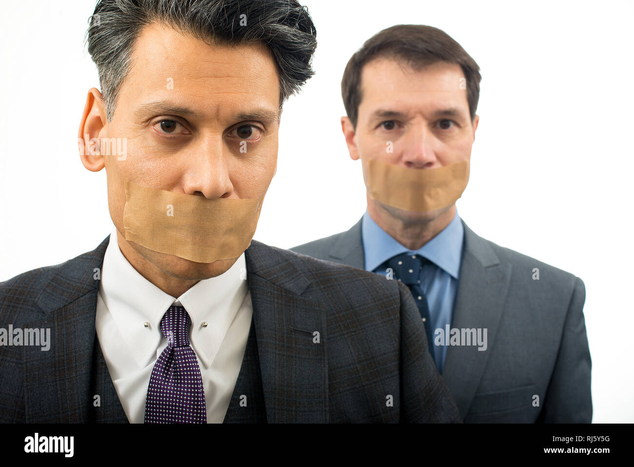 Due uomini d'affari che guardano lo spettatore in piedi contro uno sfondo bianco semplice. Entrambi gli uomini hanno la bocca imbavagliata con nastro marrone. Foto Stock