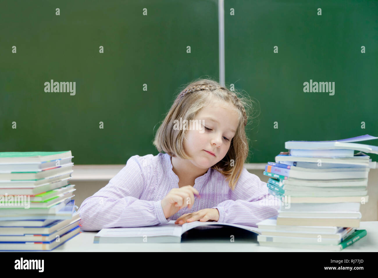Schülerin, 7 Jahre alt, lesend, zwischen zwei Stapeln Bücher, vor einer Tafel Foto Stock