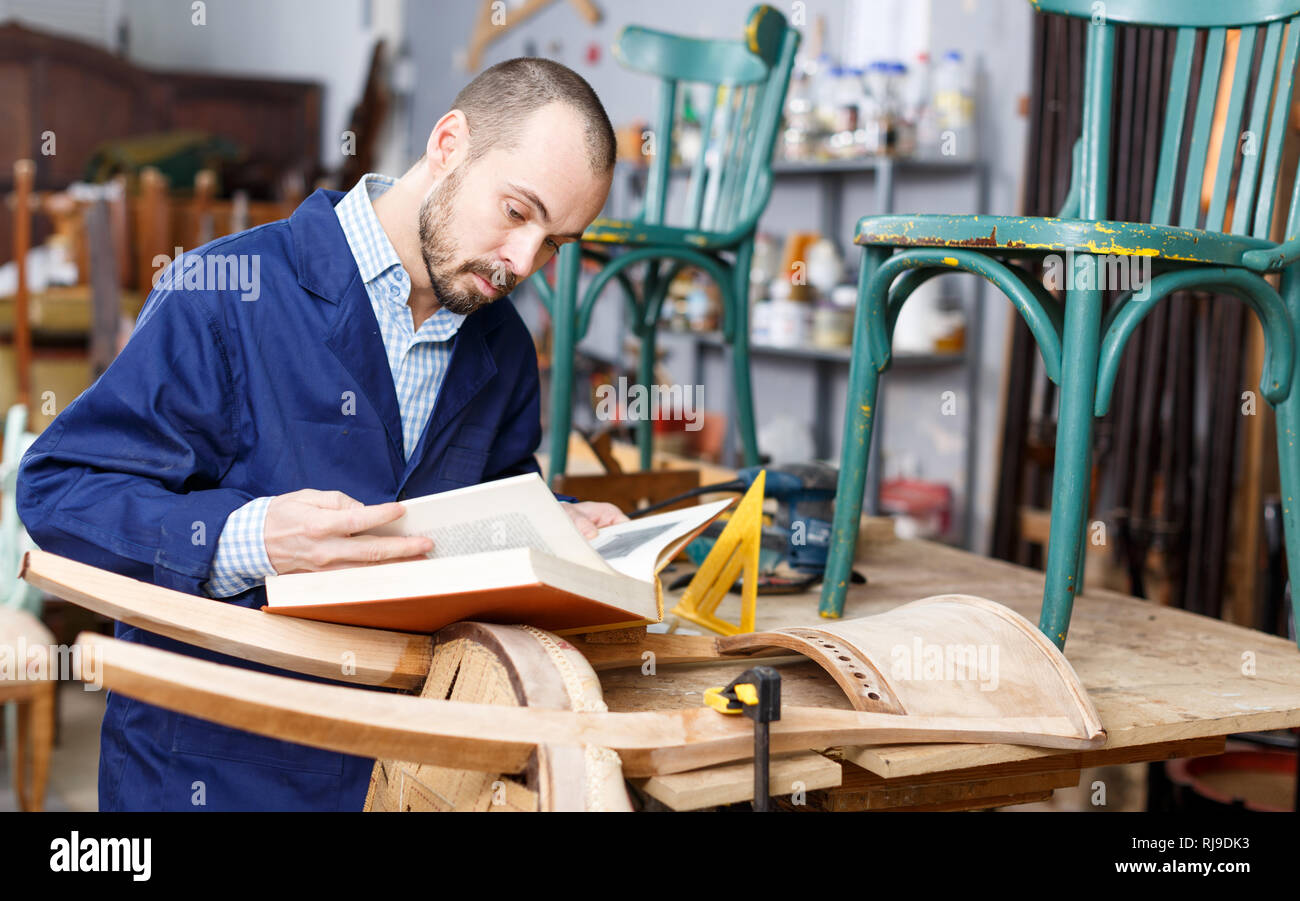 Fiducioso giovane artigiano guardando prenota prima di avviare rinnovare sedia antica Foto Stock