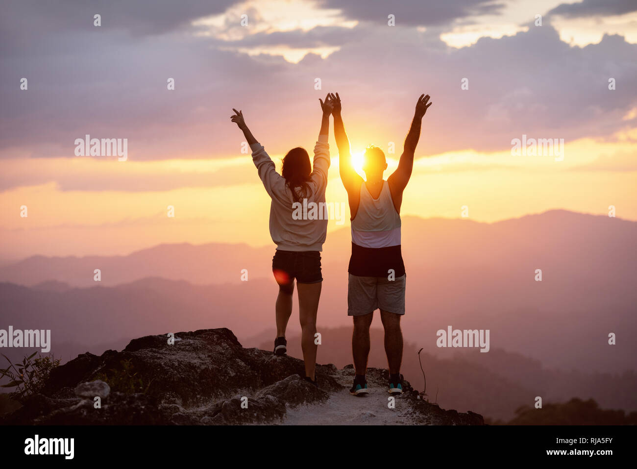 Coppia felice con i bracci sollevati sorge sulla cima della montagna contro il tramonto e divertirsi in vincitore pongono Foto Stock