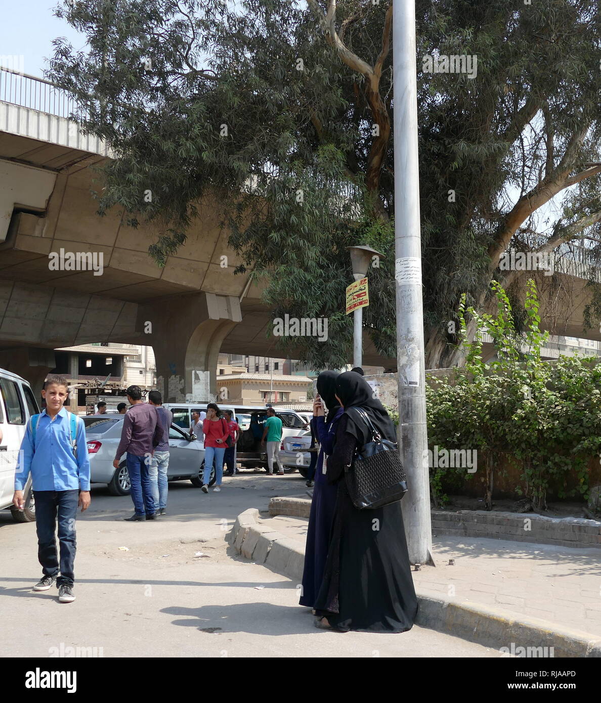 La vita quotidiana del Cairo in Egitto. Uno studente lascia la scuola come due vestita di nero donne attendere per il trasporto vicino a una strada trafficata intersezione Foto Stock