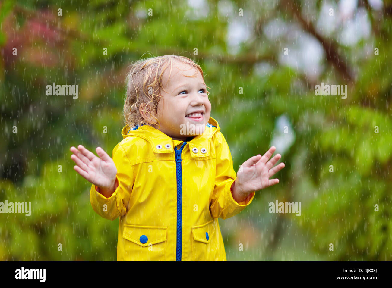 Bambini giocano in autunno la pioggia. Bambino che gioca all'esterno sul giorno di pioggia. Little Boy cattura le gocce di pioggia pesante sotto la doccia. Caduta tempesta in un parco. Usura impermeabile Foto Stock