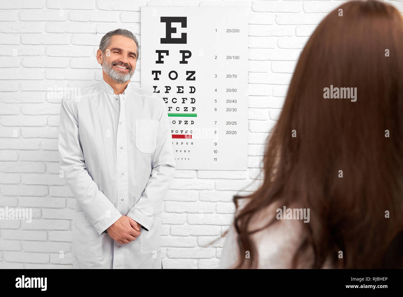 Felice medico sorridente, guardando il paziente in piedi vicino il test grafico oculare. Ophtalmologist esame e diagnosi di occhi di donna. Dottore in posa vicino al bianco muro di mattoni. Foto Stock