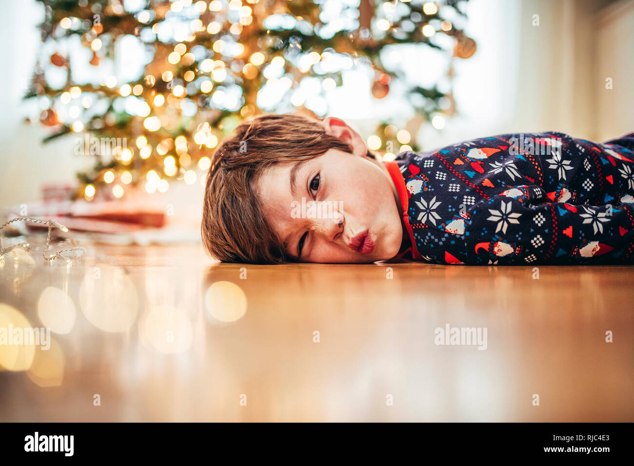 Ragazzo disteso sul pavimento nella parte anteriore di un albero di Natale tirando funny faces Foto Stock